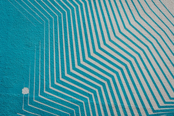 Geometric mural with diagonal white lines on a turquoise background, creating a layered, textured pattern.