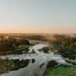 Sunset over a river with a waterfall, surrounded by lush green trees and mist rising above the water.