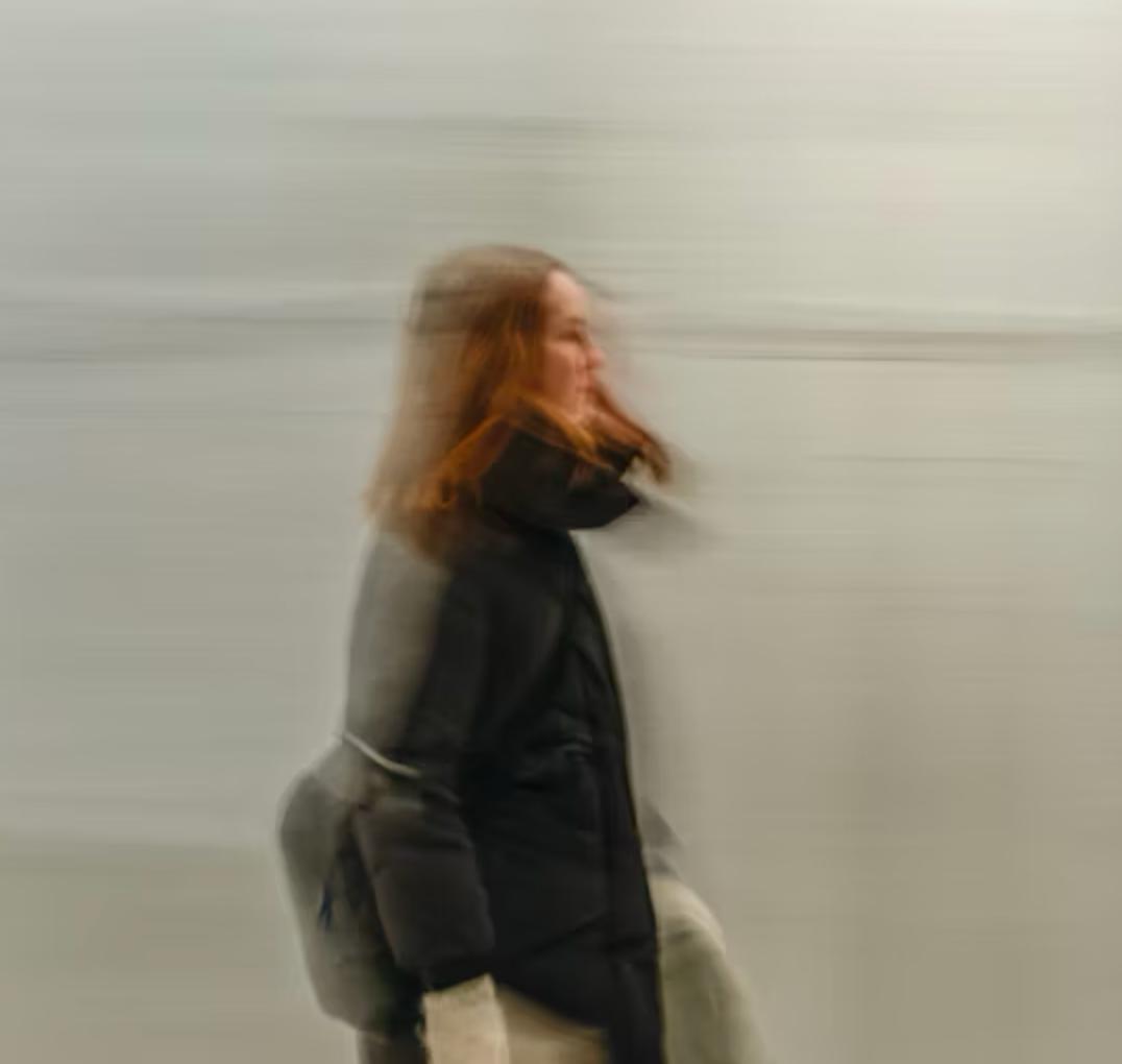 A woman with long, wavy brown hair walks briskly, wearing a black coat and white gloves, against a blurred gray background.