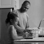 A father and daughter wash their hands at a kitchen sink, with a dish rack in the background. The scene is in black and white.