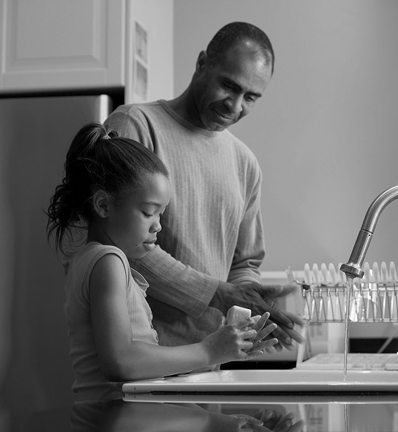 A father and daughter wash their hands at a kitchen sink, with a dish rack in the background. The scene is in black and white.