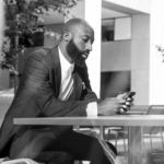Man with a beard sitting at a table outdoors, looking at his phone. Modern building and greenery in the background.