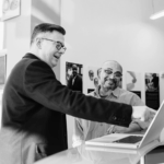 Two men engage in conversation while looking at a laptop, smiling. The setting is bright with posters in the background.