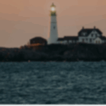 Lighthouse on rocky shore at sunset, with water in the foreground and a soft glow from the light atop the tower.