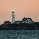 White lighthouse with green light stands on rocky shore at sunset, next to a white house, with calm ocean in foreground.