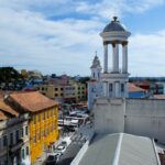 View of a vibrant cityscape with colorful buildings, a clock tower, and a market area under a cloudy sky.
