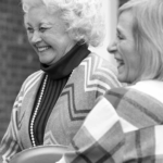 Two women laughing and smiling; one has curly white hair and wears a patterned sweater, the other has straight blonde hair and wears a plaid shirt.