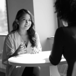 Two women are seated at a round table in a well-lit room. One woman, with long hair and wearing a light-colored top, is speaking animatedly, while the other woman, facing away, is wearing a dark top and listening attentively.
