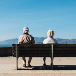 Two elderly people sit on a bench, facing away, overlooking a scenic view of mountains and water under a clear blue sky.