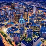 Aerial view of a city skyline at night, featuring illuminated skyscrapers and bridges over rivers, with a vibrant urban landscape.