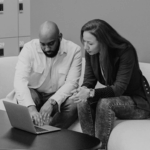 Two people, a man and a woman, are sitting on a light-colored couch, focused on a laptop on a black table. The man has a bald head and a beard, wearing a white shirt. The woman has long hair and is wearing a dark blazer over a polka dot shirt. They appear engaged in discussion.