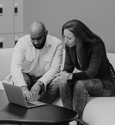 Two people, a man and a woman, are sitting on a light-colored couch, focused on a laptop on a black table. The man has a bald head and a beard, wearing a white shirt. The woman has long hair and is wearing a dark blazer over a polka dot shirt. They appear engaged in discussion.