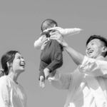 A man joyfully lifts a baby above his head while a woman smiles at them, set against a clear sky.