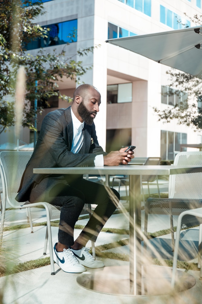 A man with a beard sits at a metal table in an outdoor area, using his phone. He wears a dark blazer, white shirt, and sneakers.