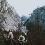 Two hikers with backpacks stand on a rocky outcrop, looking at a valley below with trees and a waterfall, one points towards the view.