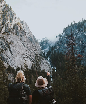 Two hikers with backpacks stand on a rocky outcrop, looking at a valley below with trees and a waterfall, one points towards the view.