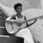 Smiling young woman with short curly hair playing an acoustic guitar while sitting on stone steps.