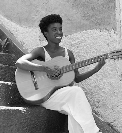 Smiling young woman with short curly hair playing an acoustic guitar while sitting on stone steps.