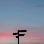 Silhouette of a directional signpost against a colorful sunset sky with pink and blue hues.
