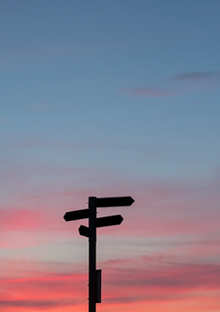 Silhouette of a directional signpost against a colorful sunset sky with pink and blue hues.