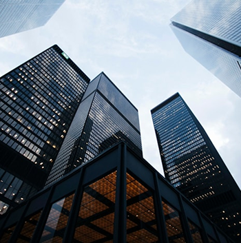Skyscrapers viewed from below, featuring glass facades and a cloudy sky above. The lower structure has warm-toned lighting.