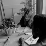 Two people sketching at a table with notebooks, while a woman gestures and discusses ideas. A potted plant is visible.