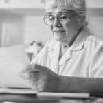 Elderly woman with glasses reading a document, seated at a table with a focused expression. Black and white image.