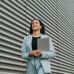 Smiling woman in a light blue suit holding a laptop, walking beside a striped metal wall.