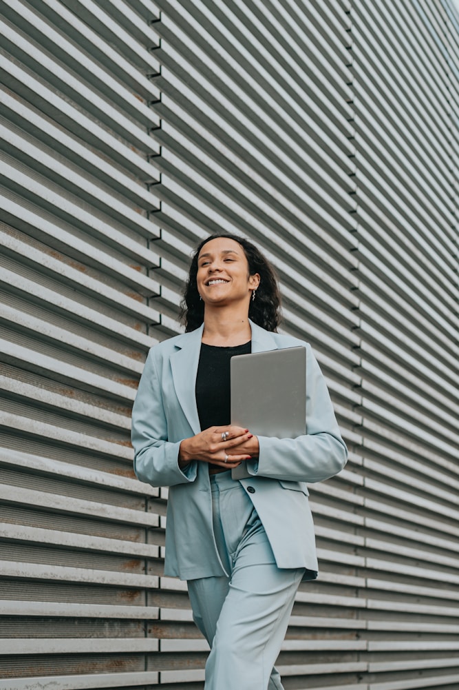 Smiling woman in a light blue suit holding a laptop, walking beside a striped metal wall.