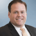 Smiling man in a dark suit and light orange tie, with short brown hair, against a light blue background.