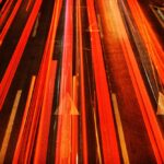 Bright red and orange light streaks from moving vehicles on a dark road with white arrow markings indicating direction.