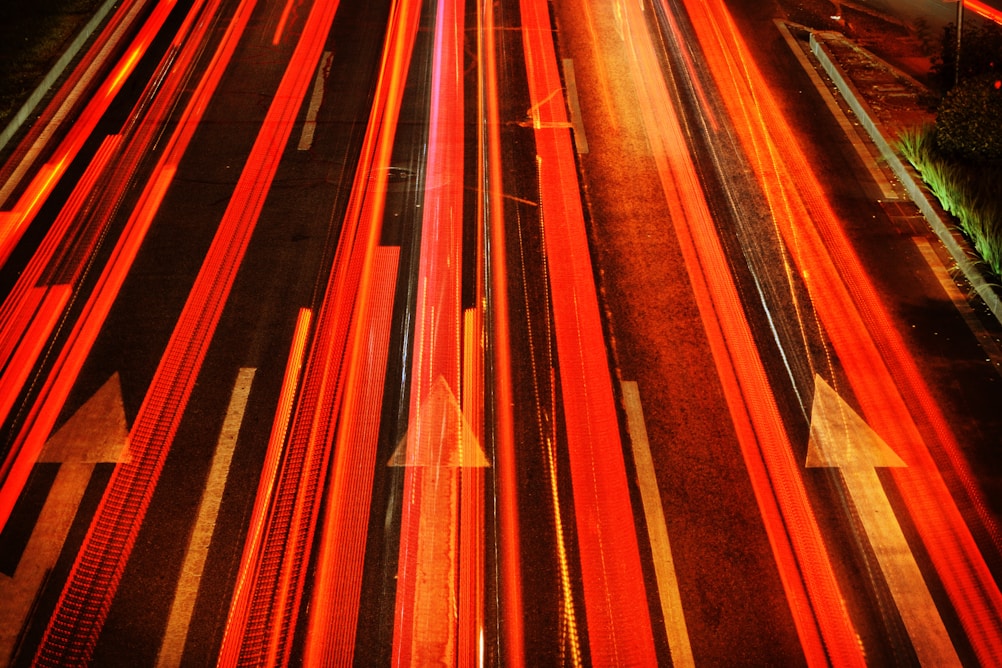 Bright red and orange light streaks from moving vehicles on a dark road with white arrow markings indicating direction.