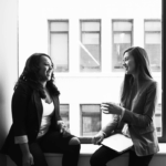 Two women sitting by a window, smiling and engaging in conversation. One woman has curly hair and is wearing a black blazer, while the other has straight hair and is in a gray blazer.