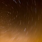 Star trails create circular patterns against a dark sky, with a warm orange glow at the bottom, suggesting a horizon.