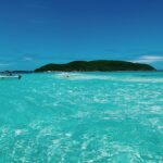 Turquoise waters with gentle waves in the foreground, an island with greenery in the background under a clear blue sky.