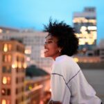 Smiling woman with curly hair leans on a rooftop railing, overlooking a city skyline at dusk with buildings illuminated.