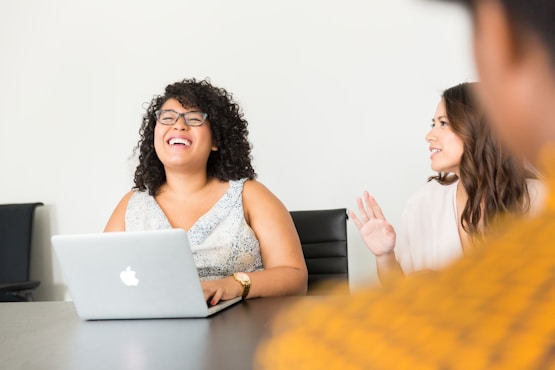 A woman with curly hair and glasses laughs while seated at a table with a laptop. Another woman gestures with her hand, smiling.