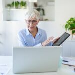 Elderly woman with short gray hair and glasses sits at a desk, holding a tablet and gesturing while looking at a laptop. Green plants are in the background.