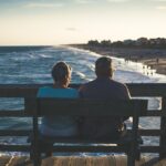 Elderly couple sitting on a wooden bench, overlooking a beach at sunset, with waves and people walking along the shore.
