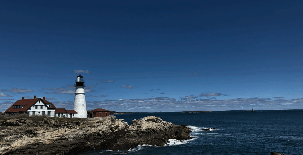 Lighthouse with a white tower and red-roofed building on rocky coast, under a blue sky with scattered clouds, ocean waves visible.