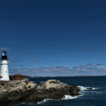Lighthouse with a white tower and red-roofed building on rocky coast, under a blue sky with scattered clouds, ocean waves visible.