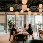 Four women sit around a wooden table in a modern office, sharing a meal. Soft paper lanterns hang from the ceiling, and large windows provide a view of the city.