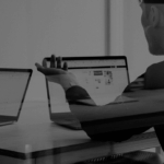 Two people sit at a table with laptops, discussing something. One person gestures with their hand, while the other types.