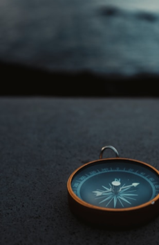 A close-up of a vintage compass with a dark blue dial and orange rim, resting on a textured gray surface, with a blurred background.