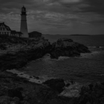 A rocky coastline features a tall lighthouse beside a dark house, under a cloudy sky, with waves crashing against the rocks.