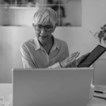 Senior woman with short gray hair and glasses, smiling while holding a tablet and gesturing in front of a laptop.
