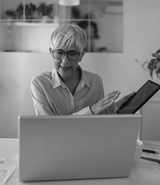 Senior woman with short gray hair and glasses, smiling while holding a tablet and gesturing in front of a laptop.