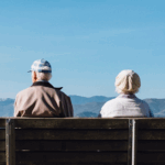 Two elderly individuals sit on a wooden bench, facing away, with mountains and a clear blue sky in the background.