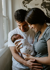 A smiling couple holds a newborn wrapped in a star-patterned blanket, standing by a window with a world map in the background.