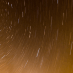 Long exposure photo showing a starry night sky with streaks of light creating circular patterns against a dark orange background.